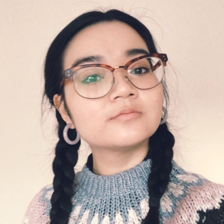 Elisa has her dark hair in two long plaits. She's looking at the camera with her head slightly tilted upwards. She's wearing a fair isle knit jumper in tones of dark blue, pink and grey, and her hoop earrings are a matching pale pink colour. She's wearing glasses that has an upper rim in tortoiseshell brown. 